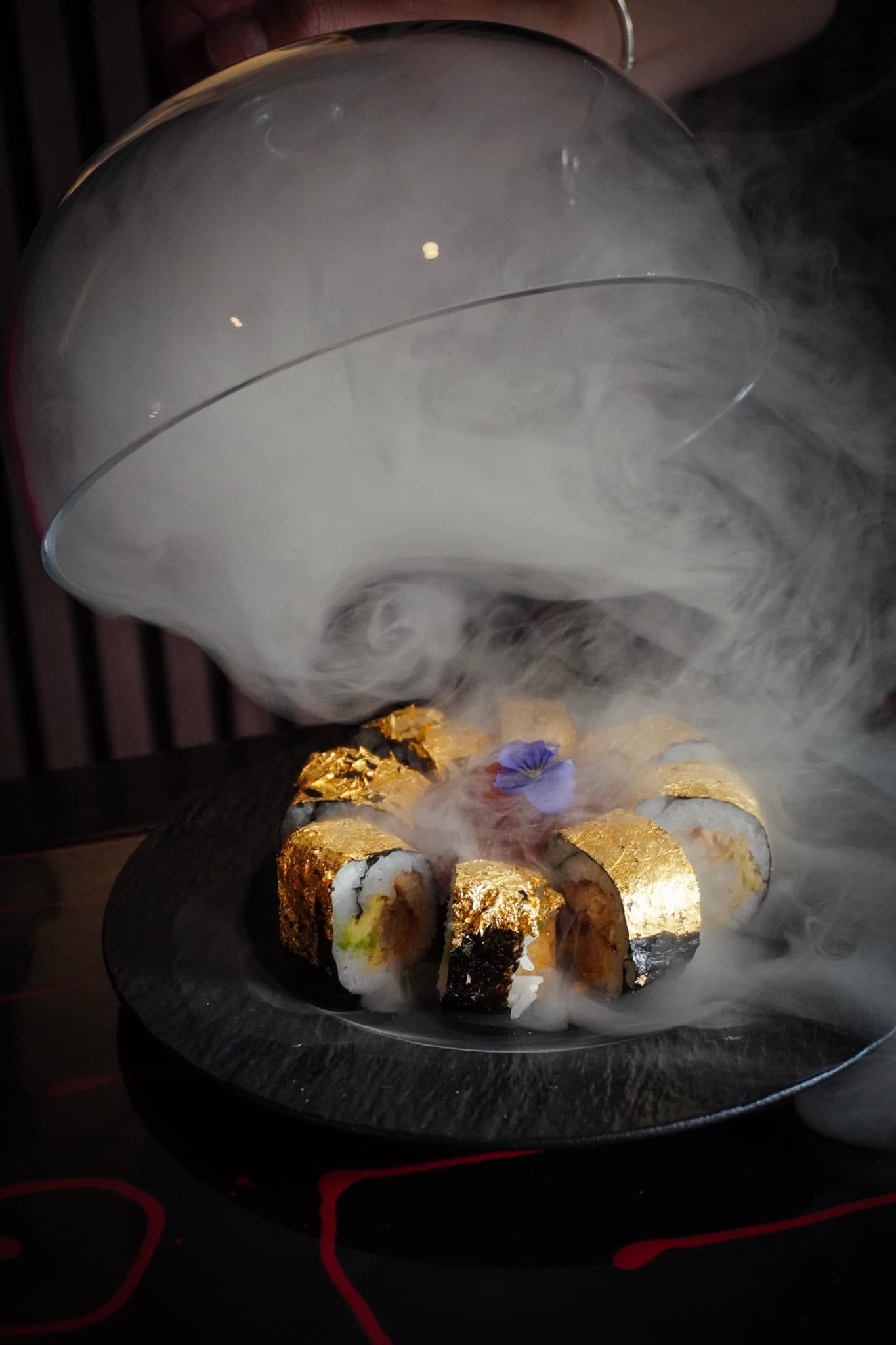 A gold leaf sushi roll revealed from under a glass dome with theatrical smoke.