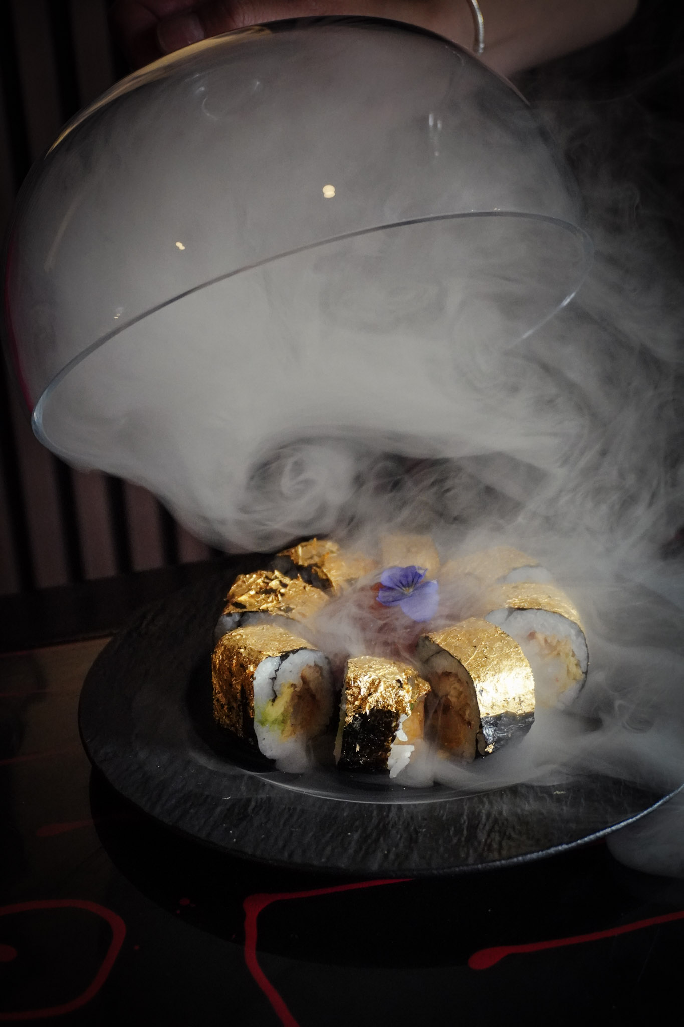 A gold leaf sushi roll revealed from under a glass dome with a burst of theatrical smoke.